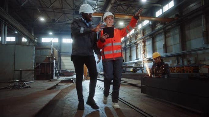 两名重工业工程师走进钢铁金属制造厂，使用数字平板电脑并进行讨论。黑人非裔美国工业专家与女技术员交谈。