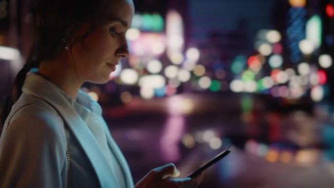美丽的年轻女子使用智能手机走过充满霓虹灯的夜晚城市街道。使用手机的华丽微笑女性肖像。侧面轮廓视图跟踪