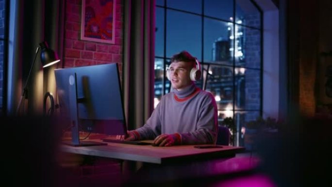 戴着耳机的年轻帅哥晚上在时尚的阁楼公寓里用电脑。有创造力的男性在家工作，浏览互联网和社交媒体。从大窗