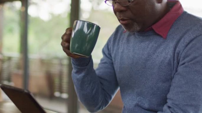 非裔美国高级男子坐在厨房的柜台上使用平板电脑并喝咖啡