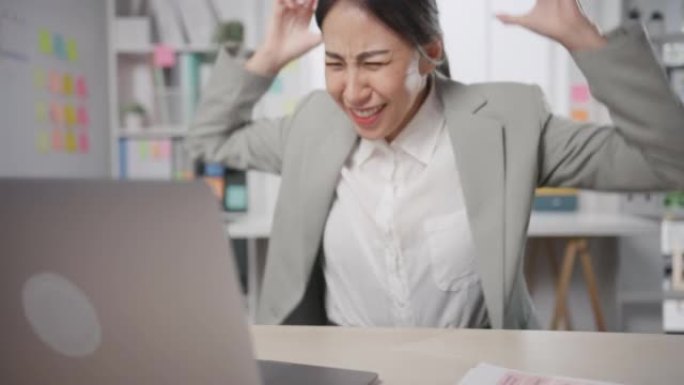 年轻的亚洲女商人坐在办公桌上疯狂地工作，压力过大，愤怒的大规模工作在办公室尖叫。