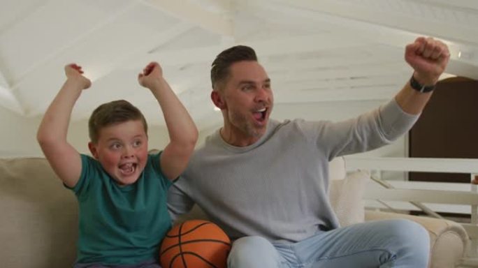 快乐的高加索父亲和儿子在客厅看电视和拿着篮球