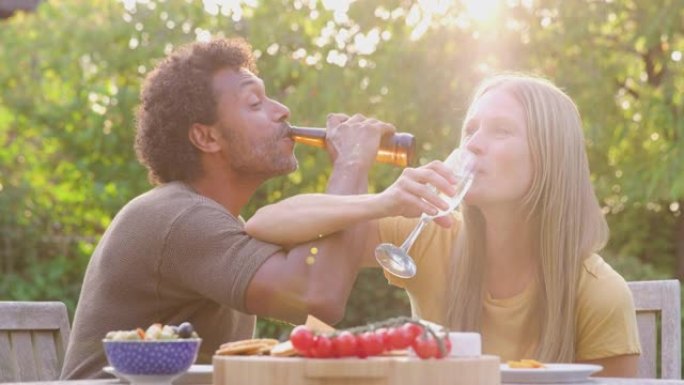 成熟的夫妇在花园里的餐桌上用香槟和啤酒和小吃庆祝时，双臂相连