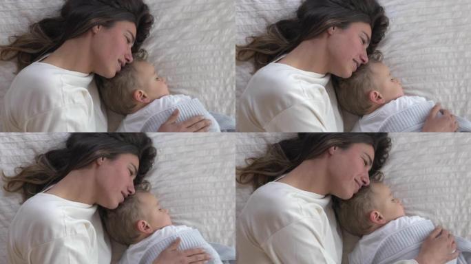 年轻母亲的真实照片是在托儿所里用柔软的毯子在床上睡觉时爱抚着她蹒跚学步的男婴。家庭、舒适、关怀、爱、
