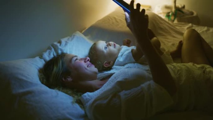 真实的快乐neo母亲和她的新生婴儿的照片在睡觉前在床上使用科技平板电脑进行家庭娱乐。