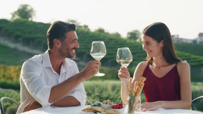 真实的爱情幸福夫妇的镜头是一起享受浪漫的晚餐，并与白酒杯欢呼，以庆祝他们的周年纪念和永恒的爱情在风景