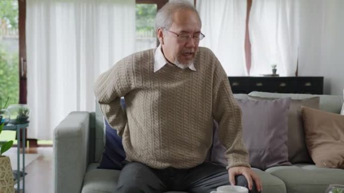 慢动作: 亚洲老年男性爷爷坐在家里客厅的沙发沙发上检疫概念保健、慢性健康问题或退休老年人的腰痛。
