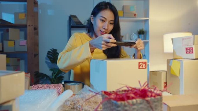 年轻的亚洲女性使用智能手机在包裹产品上拍摄条形码照片，以便晚上在家庭办公室运送给客户。