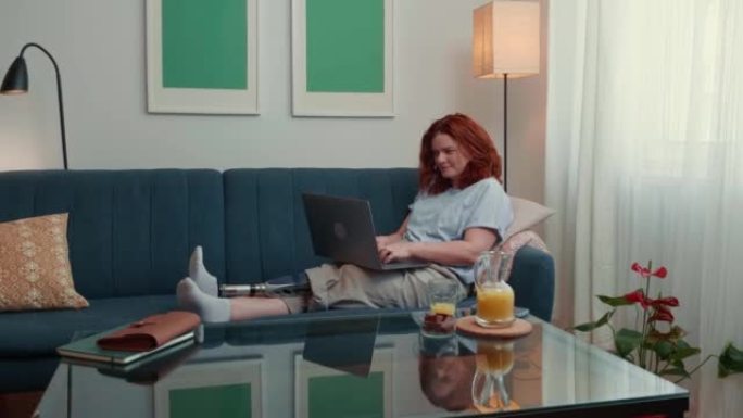 快乐的年轻美丽的高加索女商人与仿生假肢在线工作，在家里的沙发上使用笔记本电脑。