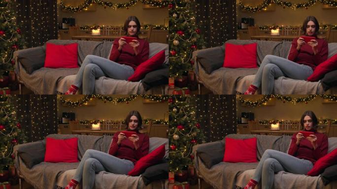 一个可爱的年轻女子正在她的红色智能手机上玩游戏，坐在房间里的沙发上，房间里有圣诞灯和装饰的圣诞树