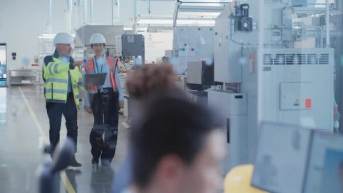 两名不同的重工业员工穿着安全制服和安全帽在工厂里散步和交谈。机架焦点切换到计算机上的非裔美国工程师绘