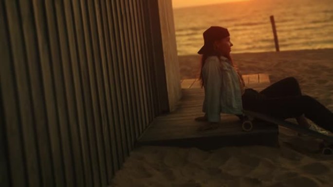 日落时，棕色头发的年轻女性坐在海滩上的木板路上，向后倾斜放松，欣赏美景