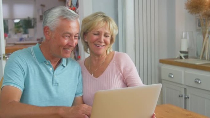资深退休夫妇坐在家里的桌子旁，用笔记本电脑进行视频通话