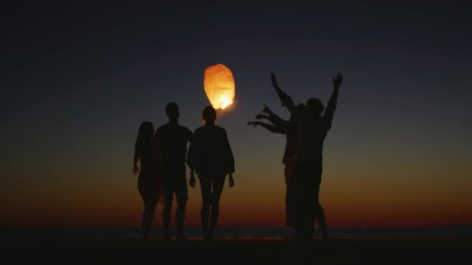 一群年轻朋友的慢动作剪影很高兴在日落时站在海滩上放下天灯。