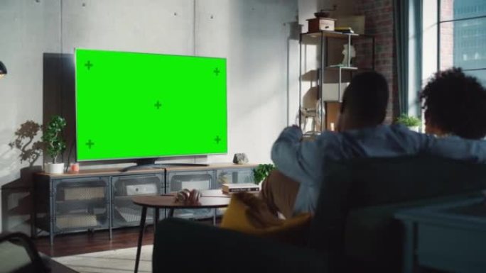 从非洲裔美国夫妇的背后拍摄，一边看电视，一边用绿屏显示，一边热烈讨论。男朋友和女朋友在家里消磨时间