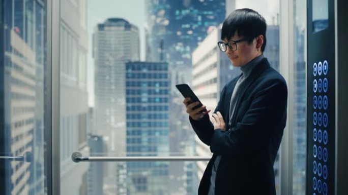 成功的时尚日本商人骑玻璃电梯到现代商务中心的办公室。英俊的男人在使用智能手机时微笑，写短信，在电梯中