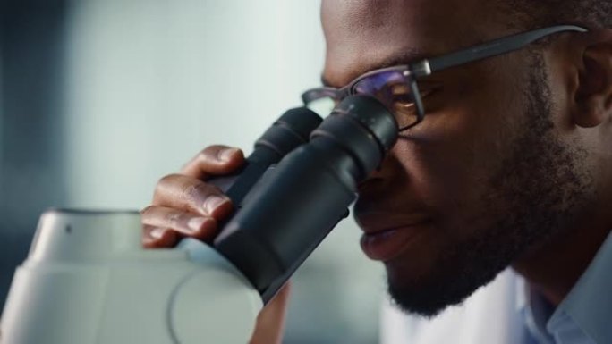 宏观特写一位英俊的黑人男性科学家戴着眼镜，看着显微镜的镜头。微生物学家使用技术设备在现代实验室中研究