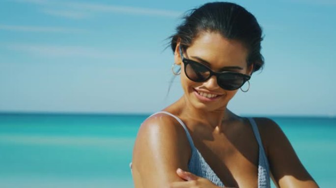 带着太阳镜的快乐年轻黑发女人的慢动作正在海滩度假期间使用防晒霜或晒黑乳液来护理她的皮肤，并在相机中微