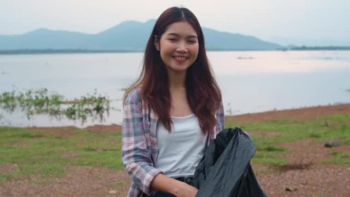 年轻的亚洲女士志愿者的肖像帮助保持大自然的清洁，在海滩上拿着塑料瓶废物和黑色垃圾袋。