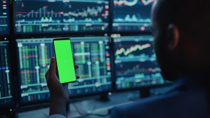 财务分析师使用带有绿屏色度键模拟模板的智能手机，并在带有股票图表的多显示器工作站上工作。商人晚上在投