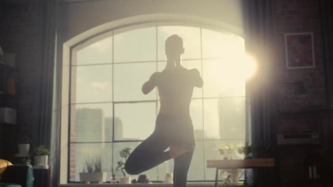 年轻的运动女子在她阳光明媚的家庭客厅锻炼身体，用Vrikshasana树瑜伽姿势练习平衡。健康的生活