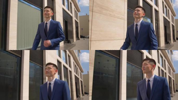 一个穿着蓝色西装的亚洲年轻人，心情愉快，他开心地笑着，在晴天去办公室上班。步行经过现代办公楼。