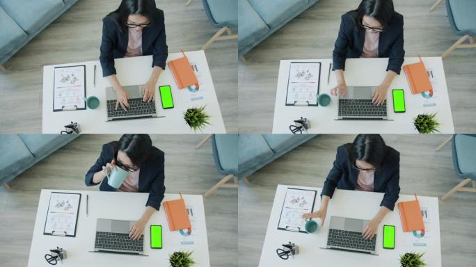 使用计算机在家工作并看着色键绿色屏幕智能手机的年轻女商人的高角度视图
