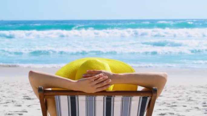 在炎热的夏日，戴着帽子的女人在海滩上放松。无法识别的白人女性躺在外面的沙滩椅上，戴着有趣的黄色遮阳帽