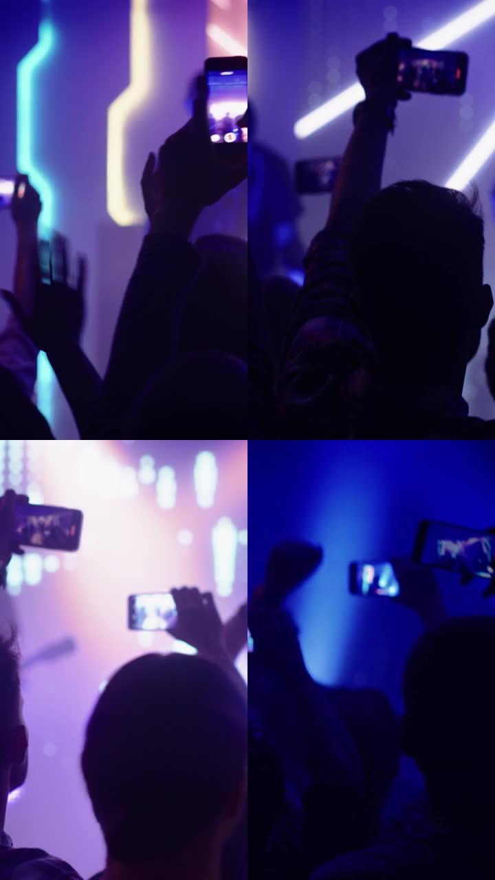 摇滚乐队在夜总会音乐会上表演的垂直镜头。前排人群在手机上录制视频。在舞台上明亮的彩色频闪灯前聚会。