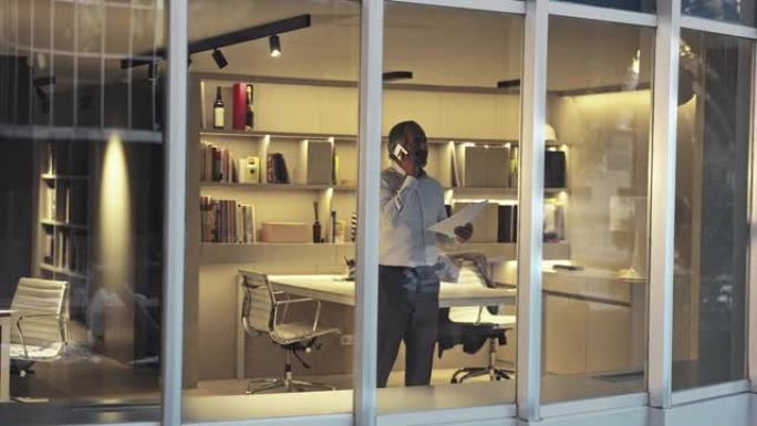 50年代中期的男性建筑师在深夜在现代办公室使用电话通过窗口观看外部视频