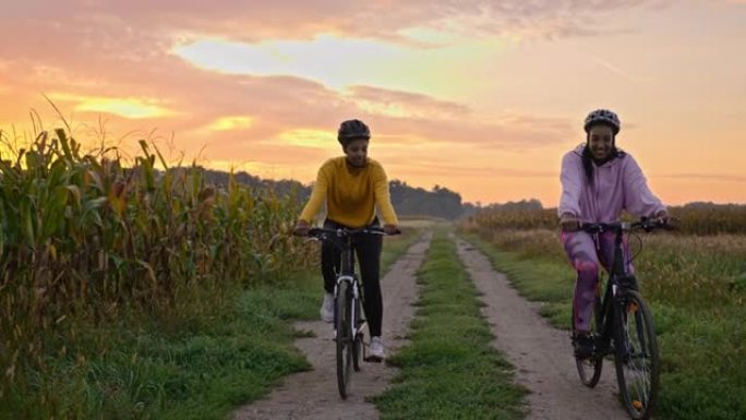跟踪两名成年成年女性在慢动作的草地土路上骑行的手持镜头，背景为日落