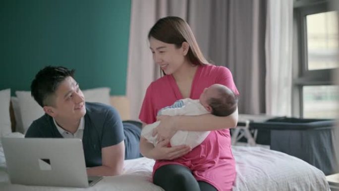 亚洲华裔父亲躺在床上使用笔记本电脑在线购物，而母亲则在卧室里抱着新生的男婴