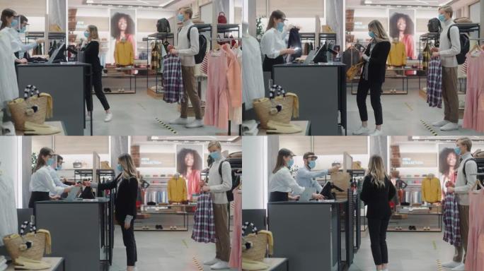 服装店: 柜台上的年轻女子从友好的零售助理那里购买衣服，并使用非接触式信用卡付款。每个人都戴着口罩。