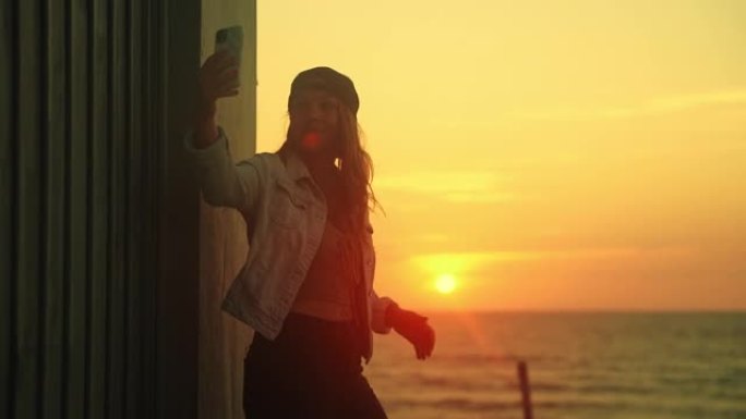 日落时，棕色头发的年轻女子站在海滩上的木板路上，背靠在建筑物上，用手机在阳光下自拍