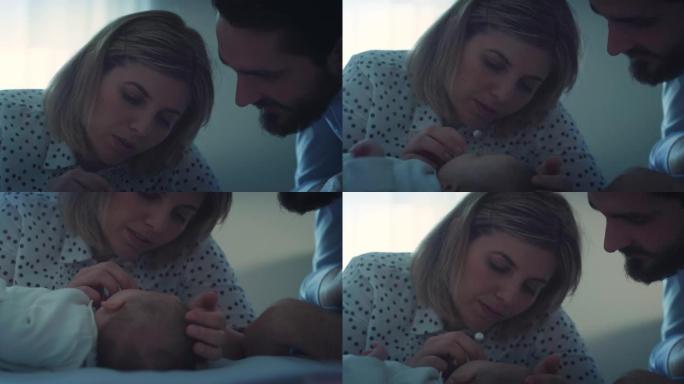 幸福家庭的电影特写镜头，新父母爱抚和亲吻他们刚出生的男婴，同时晚上在托儿所里用柔软的毯子在婴儿床里安