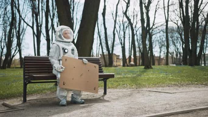 身穿宇航服的悲伤男子坐在公园的长椅上，举着车载模型标志。可怜的宇航员看着远处。穿着白色未来派西装的情