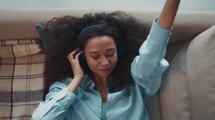 顶视图快乐美丽的混血学生女人躺在沙发上，用手机应用程序戴着耳机听音乐。