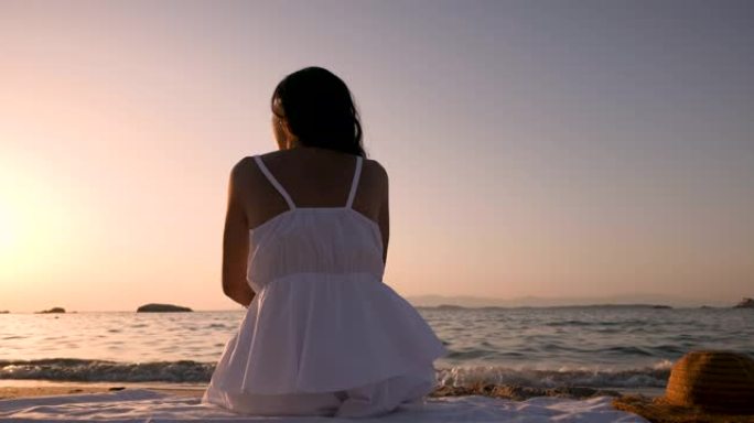 一位穿着白色太阳裙的年轻黑发女性游客坐在海边的沙滩上，在日落时分享受暑假的开始。