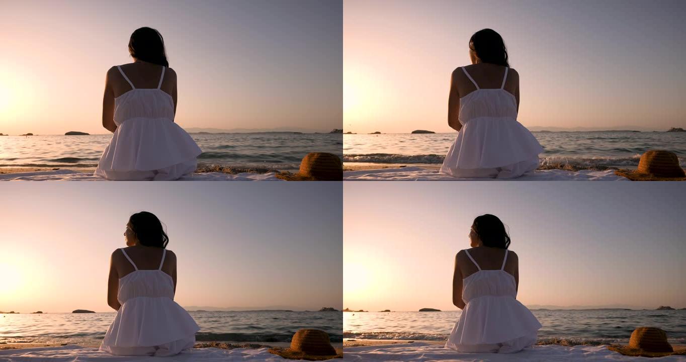 一位穿着白色太阳裙的年轻黑发女性游客坐在海边的沙滩上，在日落时分享受暑假的开始。