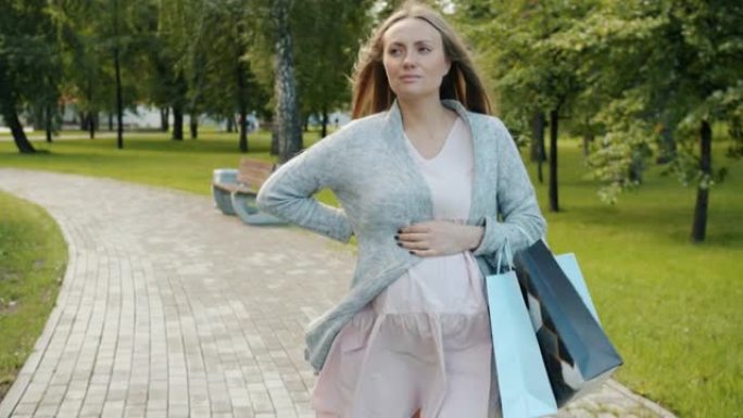 未来母亲在城市公园行走的慢动作背痛背着购物袋