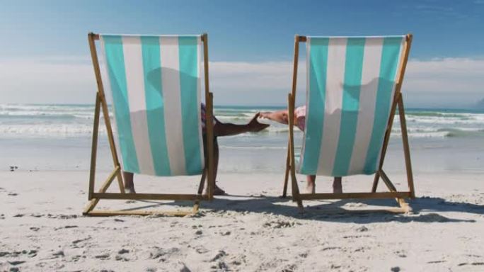 老年非裔美国夫妇坐在日光浴床上，在海滩上牵手