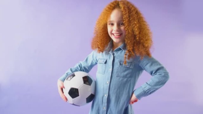 微笑的女孩在手臂下拿着足球，在紫色工作室背景下看着相机的工作室镜头-慢动作拍摄
