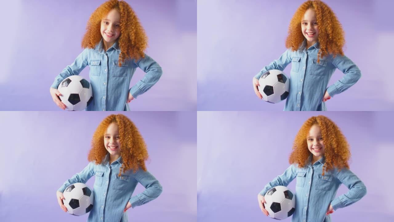 微笑的女孩在手臂下拿着足球，在紫色工作室背景下看着相机的工作室镜头-慢动作拍摄
