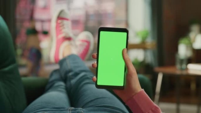 带有绿屏模拟显示的智能手机上的女性手滚动提要。女性在家里的沙发上放松，在移动设备上观看视频和阅读社交