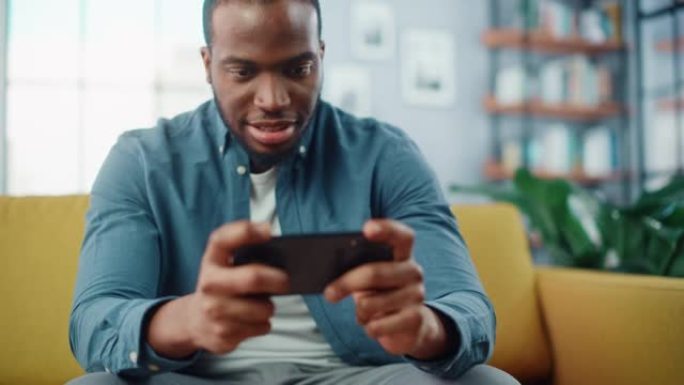 快乐的黑人非洲裔美国人坐在客厅的沙发上，在智能手机应用程序上玩视频游戏。兴奋的有色人种在家休息，在互