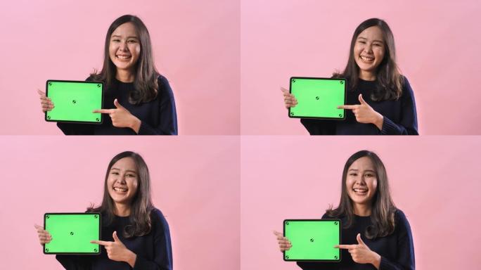 肖像亚洲妇女在粉红色的工作室背景下拿着绿色屏幕的数字平板电脑