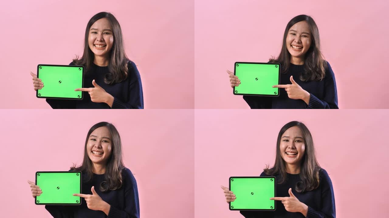 肖像亚洲妇女在粉红色的工作室背景下拿着绿色屏幕的数字平板电脑