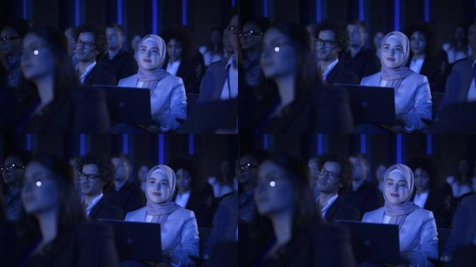 阿拉伯女性坐在科技会议上黑暗拥挤的礼堂里。使用笔记本电脑的年轻穆斯林妇女。观看创新技术演示的头巾专家