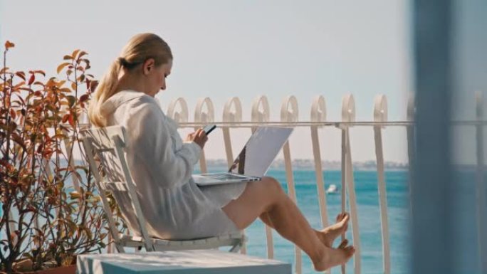 金发碧眼的女人坐在酒店房间的阳台上，在笔记本电脑和智能手机上工作，可以看到大海，前面是蓝色的窗帘