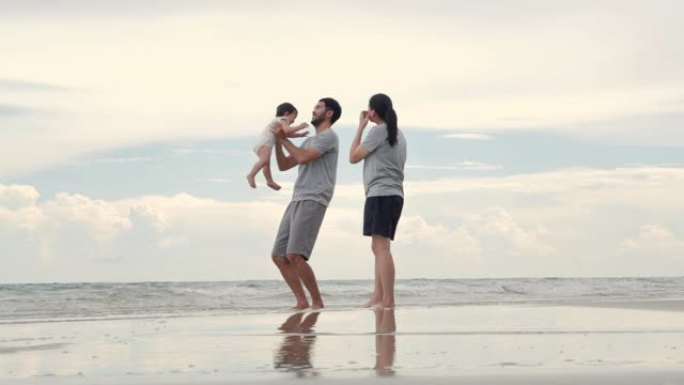 快乐的亚洲父亲，母亲，宝贝儿子在沙滩上沿着日落的边缘冲浪玩耍。活跃的父母和人们在暑假与婴儿一起户外活